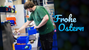 Ein Mitarbeiter der CEFEG in Chemnitz arbeitet an einem Wiegeplatz im Lager, während bunte Ostereier zwischen blauen Lagerkisten, Regalen und Bauteilen für ein Suchbild versteckt sind; rechts der Schriftzug „Frohe Ostern“.
